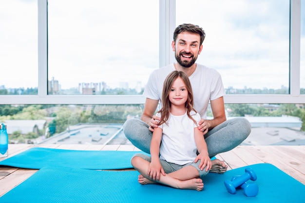 Small girl and father sitting in lotus position on yoga mats by the window