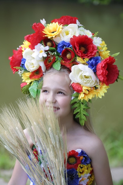 Small girl kid with long blonde hair and pretty happy smiling face in prom embriodered white dress and colorful flower national ukrainian wreath crown on head outdoor with spikelet grass