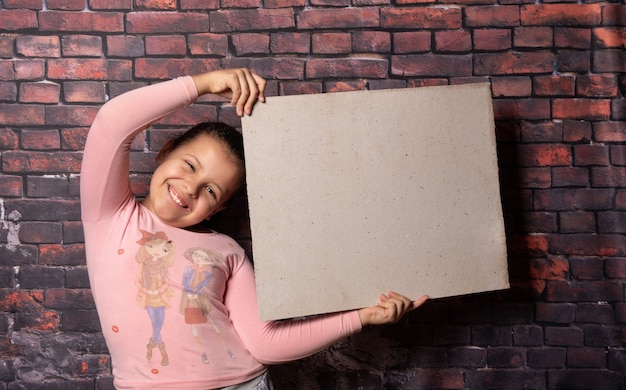 Small girl making poses with a blank recycled paper letters in front of an old brick wall backdrop, dark background, selective focus.