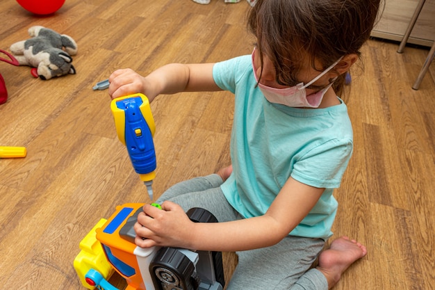 Piccola ragazza nella maschera medica che gioca con il trattore del costruttore del giocattolo a casa