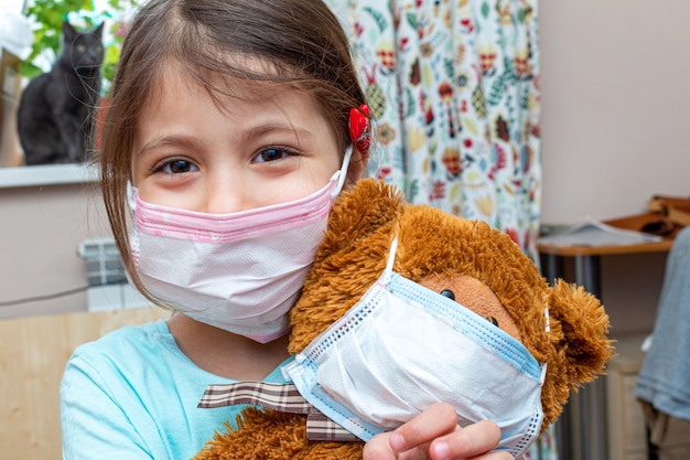 Small girl playing toy bear in medical protective mask during quarantine isolation