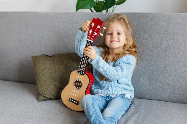 Small girl playing ukulele and singing at home. Kid learning guitar. Early childhood education