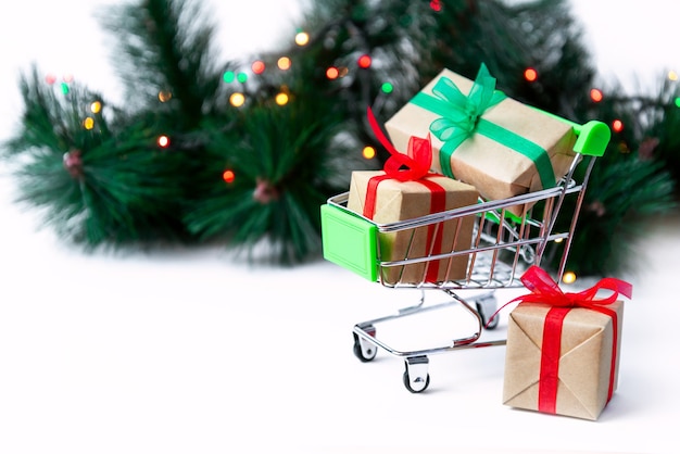 Small grocery cart with gift boxes on Christmas tree surface with garland lights