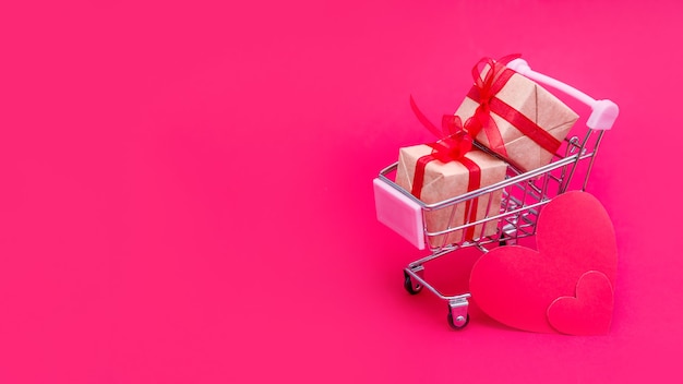 Small grocery cart with gift boxes on red-pink background. Give gifts with love on Valentine's Day,