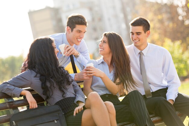 A small group of young business people during a coffee break, sitting on a park bench enjoying a coffee and a sunny day.