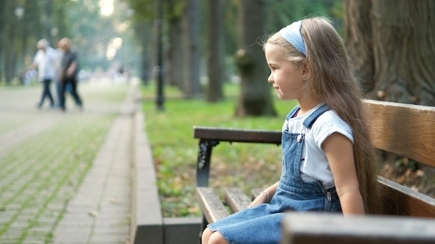 Маленькая счастливая детская девочка, сидящая на скамейке, отдыхая в летнем парке.