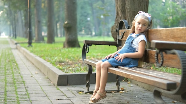 夏の公園で休んでいるベンチに座っている小さな幸せな子供の女の子