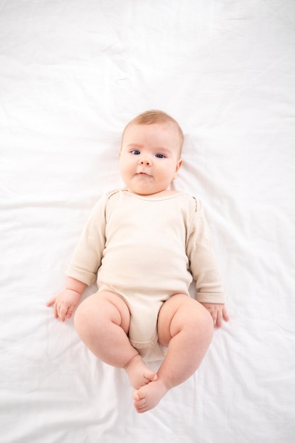 A small healthy baby in a cotton bodysuit lies on his back on a bed on white bedding in the bedroom at home looks at the camera smiles