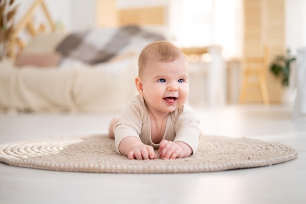 면 바디수트를 입은 작고 건강한 아기가 집의 밝은 거실에 있는 깔개 위에 엎드려 카메라 미소를 바라보고 있습니다