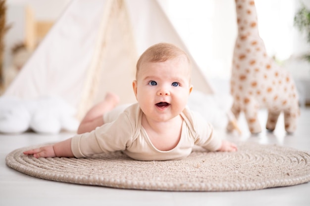 A small healthy baby in a cotton bodysuit lies on his stomach on a rug in the living room of the house against the background of a wigwam and plush toys