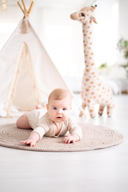 면 바디수트를 입은 작고 건강한 아기가 위그와 봉제 장난감을 배경으로 집 거실의 깔개 위에 엎드려 누워 있습니다.
