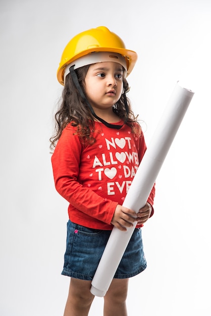 白い背景の上に分離された黄色のハード帽子と図面の紙のロールまたは計画を持つ小さなインドの女の子エンジニア