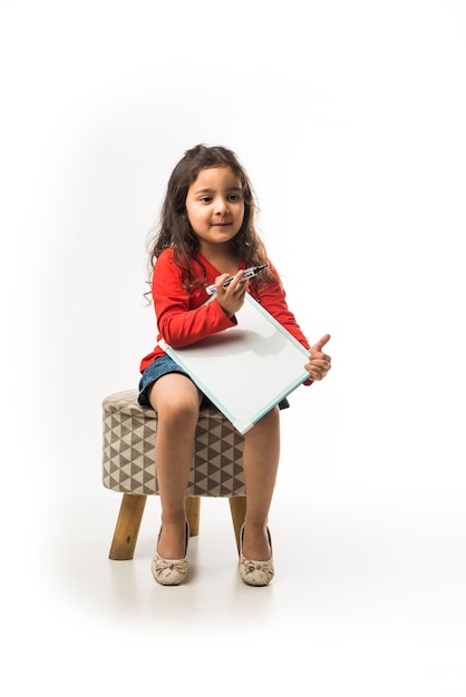 Small Indian girl writing on slate with marker pen, while sitting on stool over white background