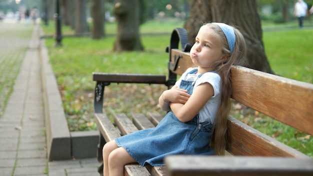 Small irritated child girl sitting alone on a bench in summer park.