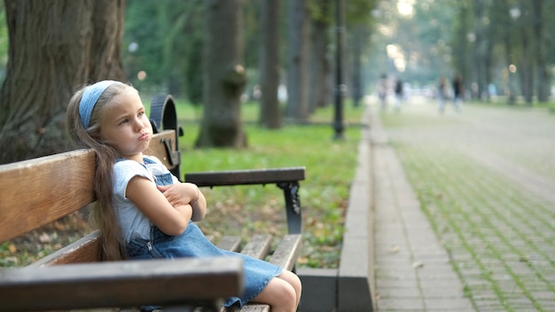 Small irritated child girl sitting alone on a bench in summer park.