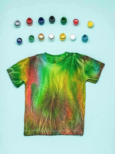 Small jars of colored fabric dyes and a tie dye T-shirt on a blue background. Flat lay.