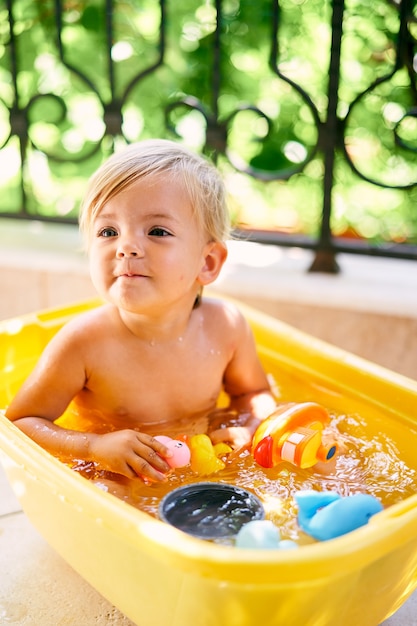 Small kid sits in a basin of water and holds rubber ducks in his hands