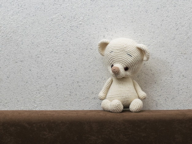 A small, lonely bear cub on the back of the sofa against the gray wall. Beautiful knitted toy.