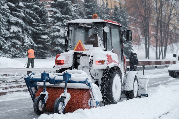 A small red tractor with a brush sweeps snow from the sidewalk
near the kremlin walls during a heavy snowfall. snowflakes are
flying in the air.