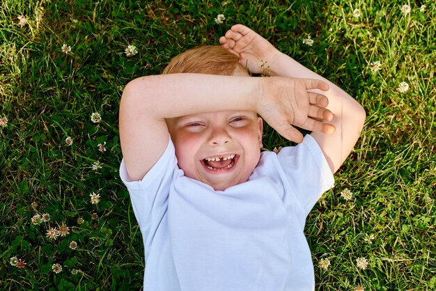 small redhaired fiveyearold boy lies on back on grass in the park and laughs top view closeup