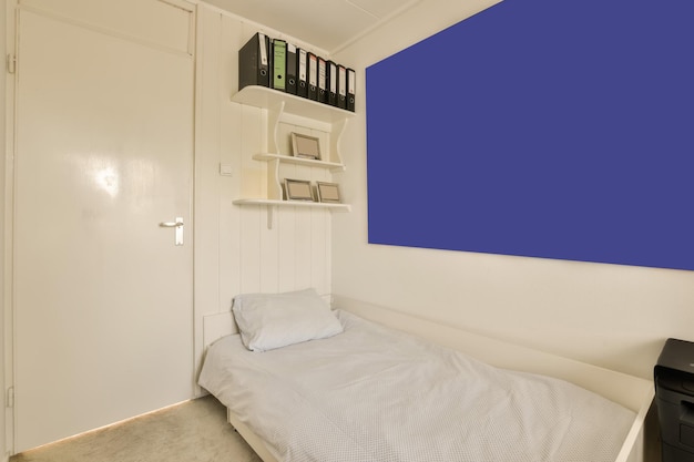 a small room with a bed desk and books on shelves above the bed is a blue wall in the background