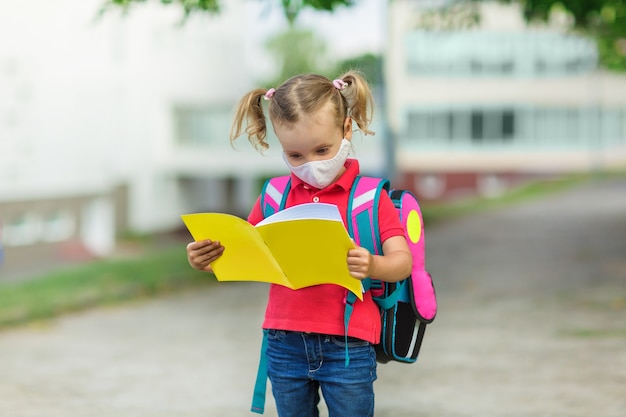 A small school girl in a medical mask comes from school with a yellow notebook in her hands