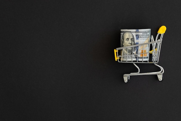 Photo small shopping cart with 100 dollar banknote on black background