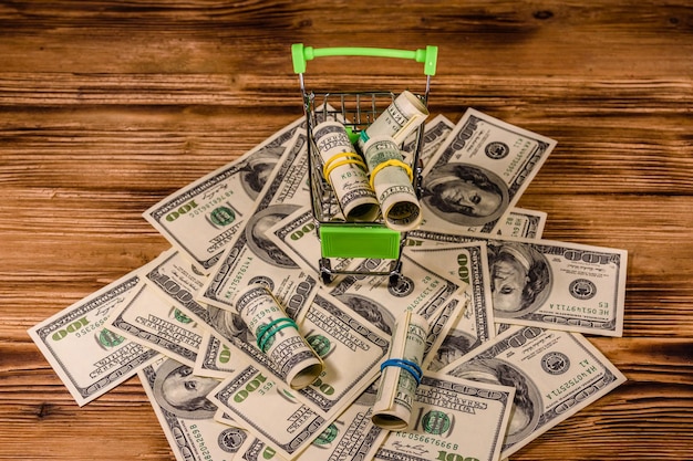 Small shopping cart with rolled up one hundred dollar banknotes on a wooden background