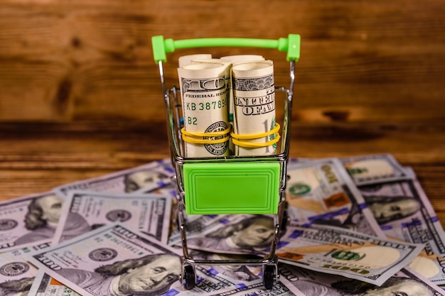 Small shopping cart with rolled up one hundred dollar banknotes on a wooden background