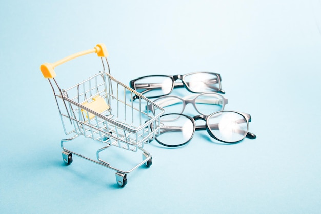 Small shopping trolley in focus, children's glasses and two pairs of black glasses for adults in blur, blue background, copy space, online purchase of glasses and eyeglass frames