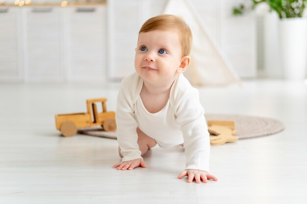 a small smiling child a boy in a white bodysuit crawls or learns to crawl on the floor of a house in a bright room with a wigwam a sixmonthold baby with blue eyes smiles or laughs