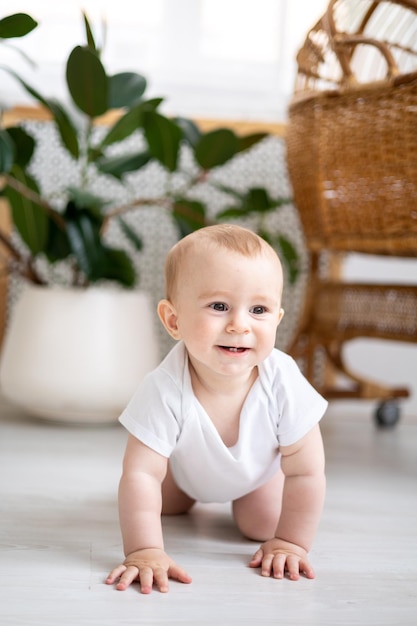 A small smiling child boy in a white bodysuit is sitting on the floor in a bright room looking at the camera smiling the child is crawling on the floor of the house