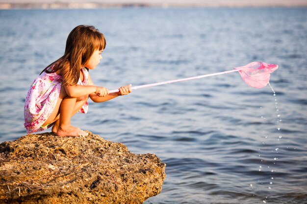 Маленькая улыбающаяся девочка в ярком купальнике стоит на солнце и смотрит на маленький камень с фоном голубого неба