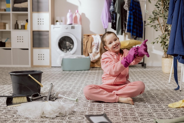 笑顔の小さな女の子が両親の床のモップ掛けを手伝っている 子供は洗濯室のバスルームに横たわり、ゴム手袋をはめた布で汚れをこすり落とす