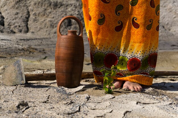 Small sprout in the desert at the feet of a woman in ethnic clothing next to a hoe and a water jug