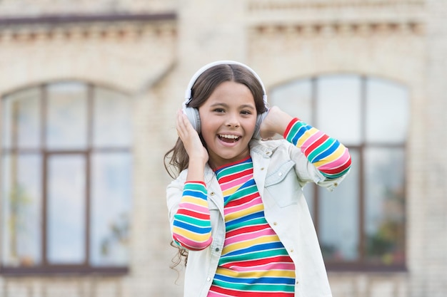 小さな学生の女の子は、学校に戻って屋外で音楽を聴きます。小さな女の子は、現代のテクノロジーを使用します。スタイリッシュなヒップスターの衣装を着た幸せな女子高生。