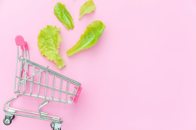 ピンクの背景に分離された緑のレタスの葉と買い物のための小さなスーパーマーケットの食料品の押しカート