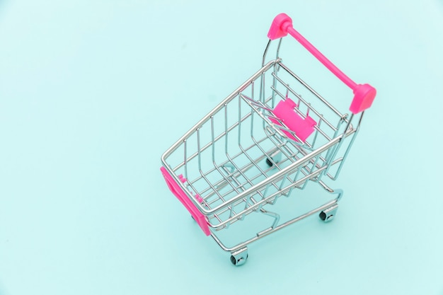 Small supermarket grocery push cart for shopping toy with wheels isolated on blue pastel colorful trendy background Copy space. Sale buy mall market shop consumer concept.