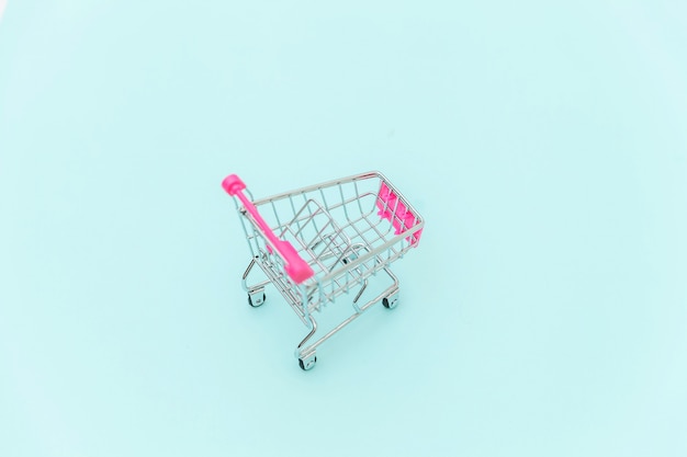 Small supermarket grocery push cart for shopping toy with wheels isolated on blue pastel colorful trendy background Copy space. Sale buy mall market shop consumer concept.