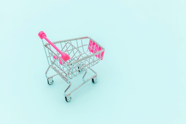 Small supermarket grocery push cart for shopping toy with wheels isolated on blue pastel colorful trendy background Copy space. Sale buy mall market shop consumer concept.