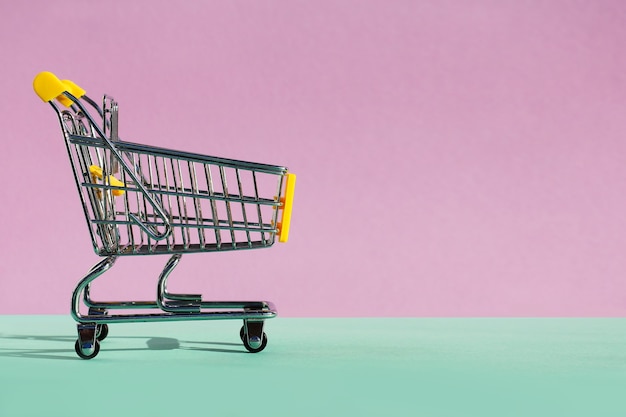 보라색-녹색 배경에 바퀴가 달린 쇼핑 장난감을 위한 작은 슈퍼마켓 식료품 푸시 카트. 소비, 매장 내 구매, 판매의 개념. 복사 공간
