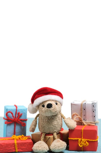 Small teddy bear in a santa claus hat sits on boxes with christmas giftson a white background