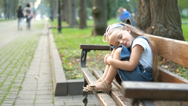 Small tired child girl sitting on a bench with closed eyes resting in summer park.