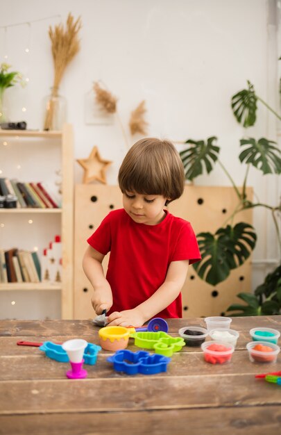 赤いTシャツを着た小さな幼児の男の子は、部屋の木製のテーブルでカラフルな粘土で遊ぶ