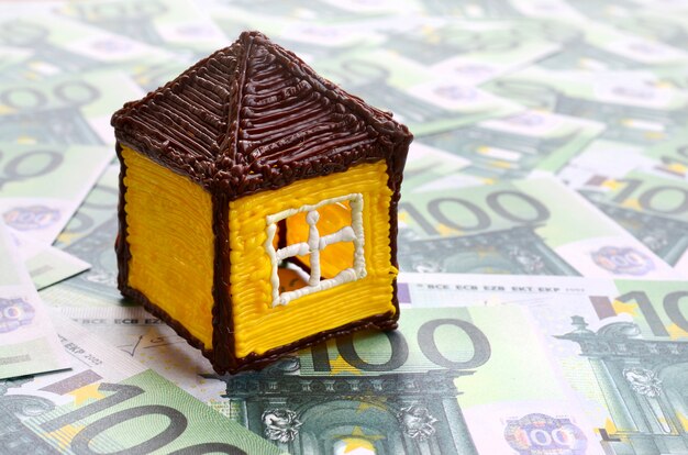 Маленький игрушечный домик лежит на множестве зеленых денежных номиналов по 100 евро