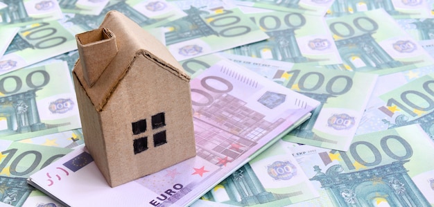 Small toy house is lies on a set of green monetary denominations of 100 euros