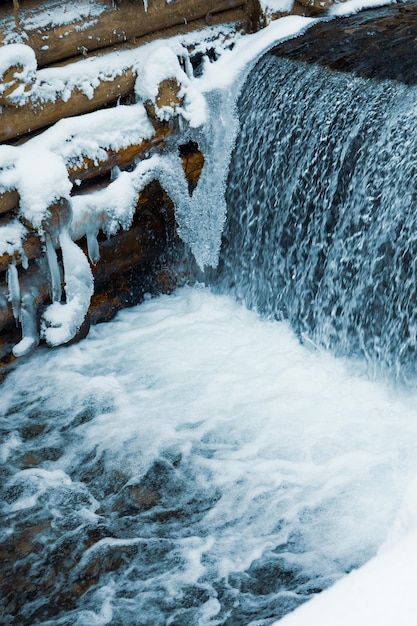 A small wall of small logs encloses a small mountain stream with waterfalls in a winter forest near bare trees in the Carpathian mountains