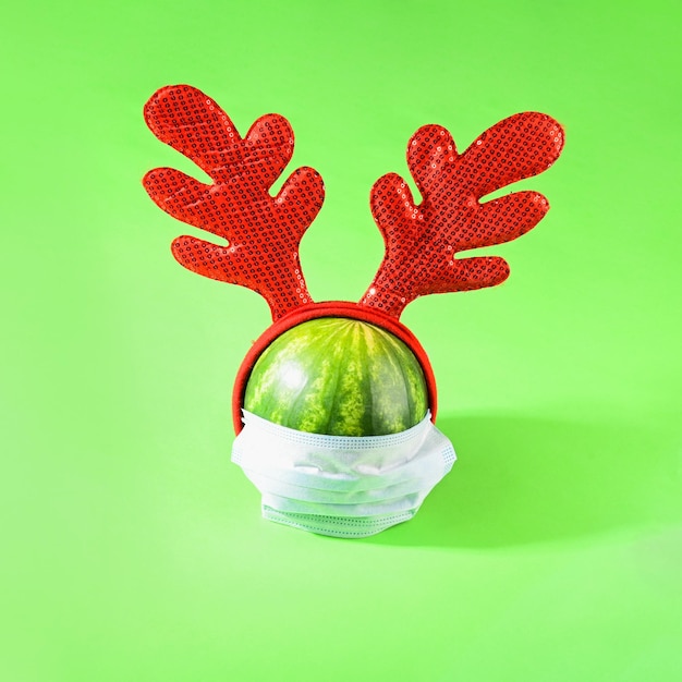 A small watermelon in a medical mask with christmas horns on a green background