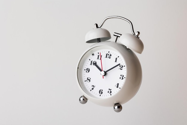 Маленький белый будильник, черные цифры, установленное время на 10.10 часов, размещенные на белом столе.