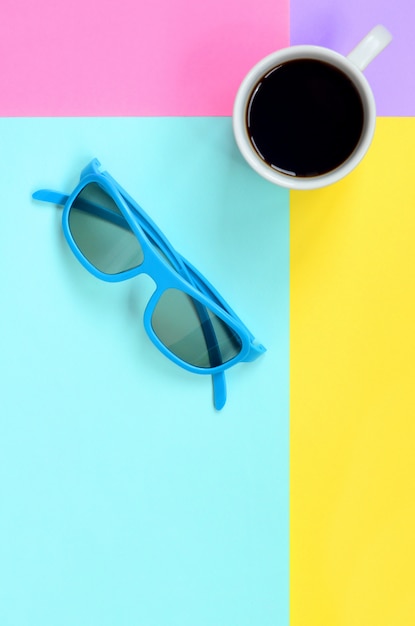 Photo small white coffee cup and blue sunglasses onfashion pastel blue, yellow, violet and pink paper